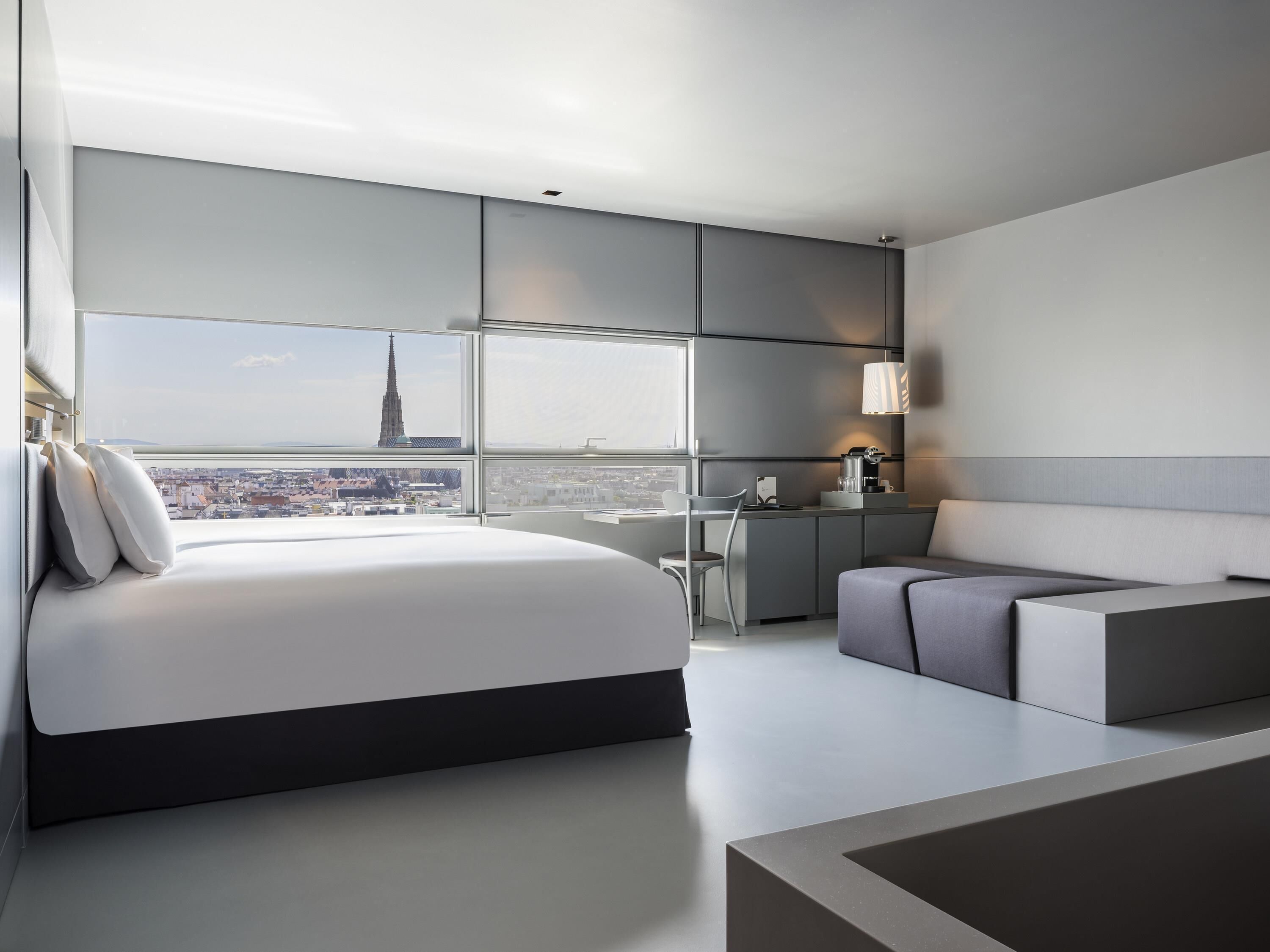 Quarto de hotel moderno com uma cama grande, área de estar, escrivaninha e vista para a cidade.