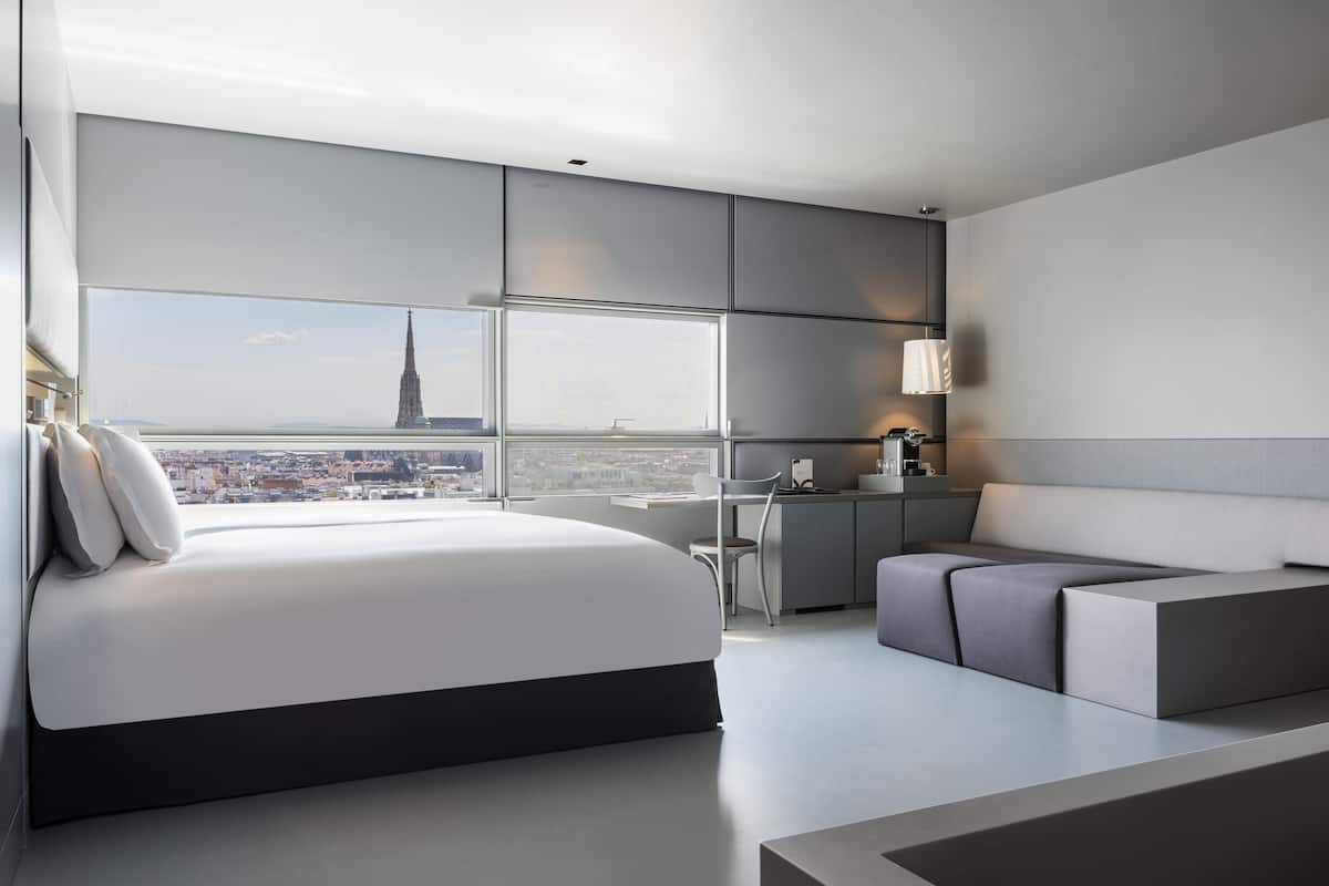 Quarto de hotel moderno com uma cama grande, área de estar, escrivaninha e vista para a cidade.