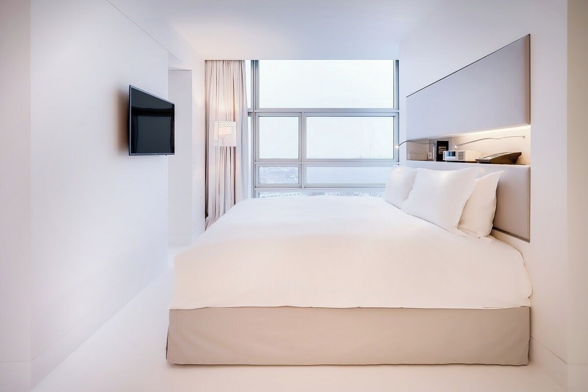 Una habitación de hotel moderna con una cama grande, un televisor de pantalla plana montado en la pared y vistas a la ciudad desde la ventana.