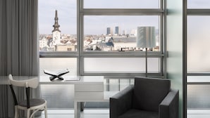 A modern room with large windows, a desk, and a comfortable chair, offering a view of a cityscape with historical and modern buildings.