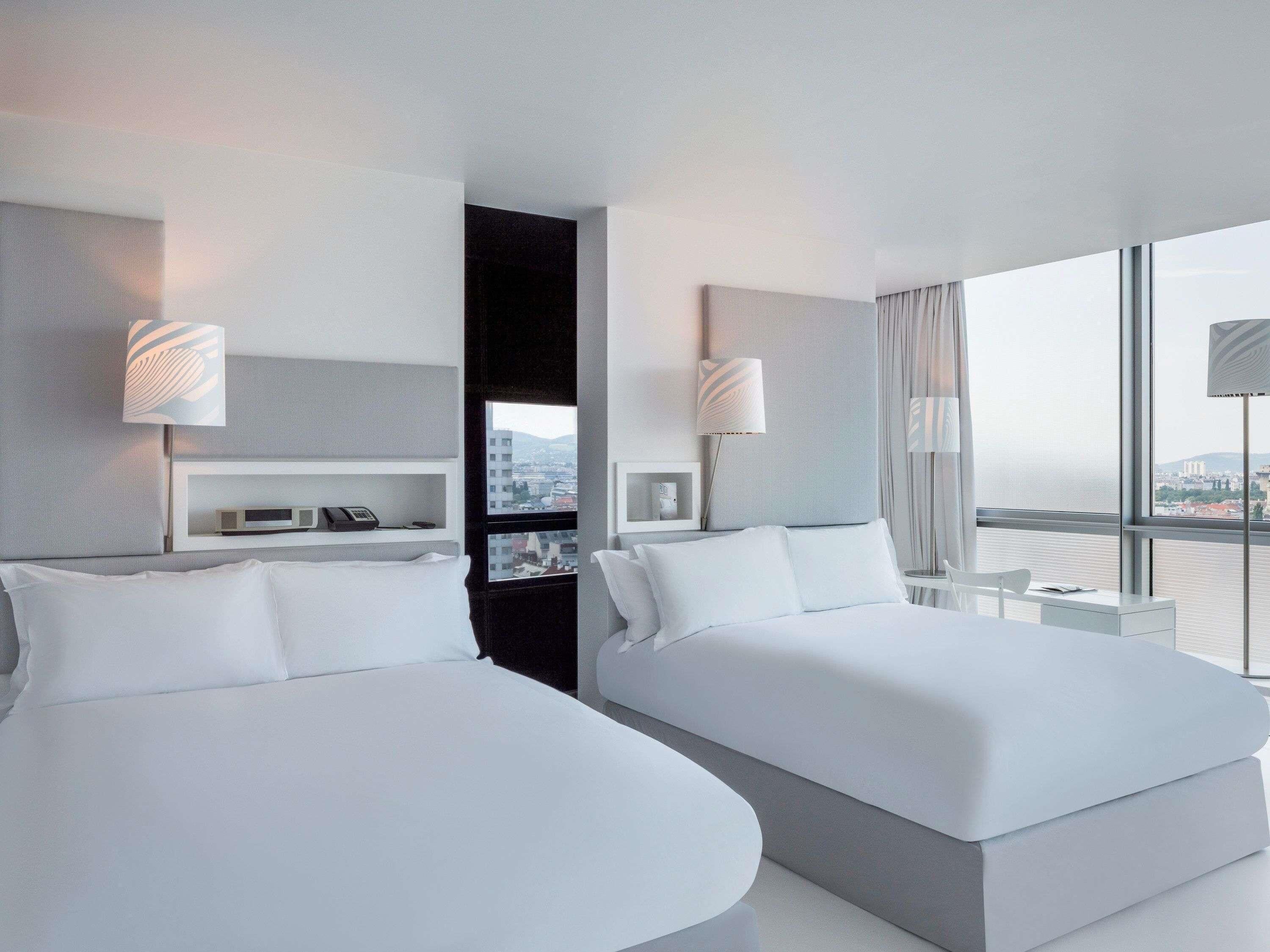 Quarto de hotel moderno com duas camas, uma pequena mesa de cabeceira, prateleira fixa com telefone e abajur, e uma grande janela com vista para a cidade.