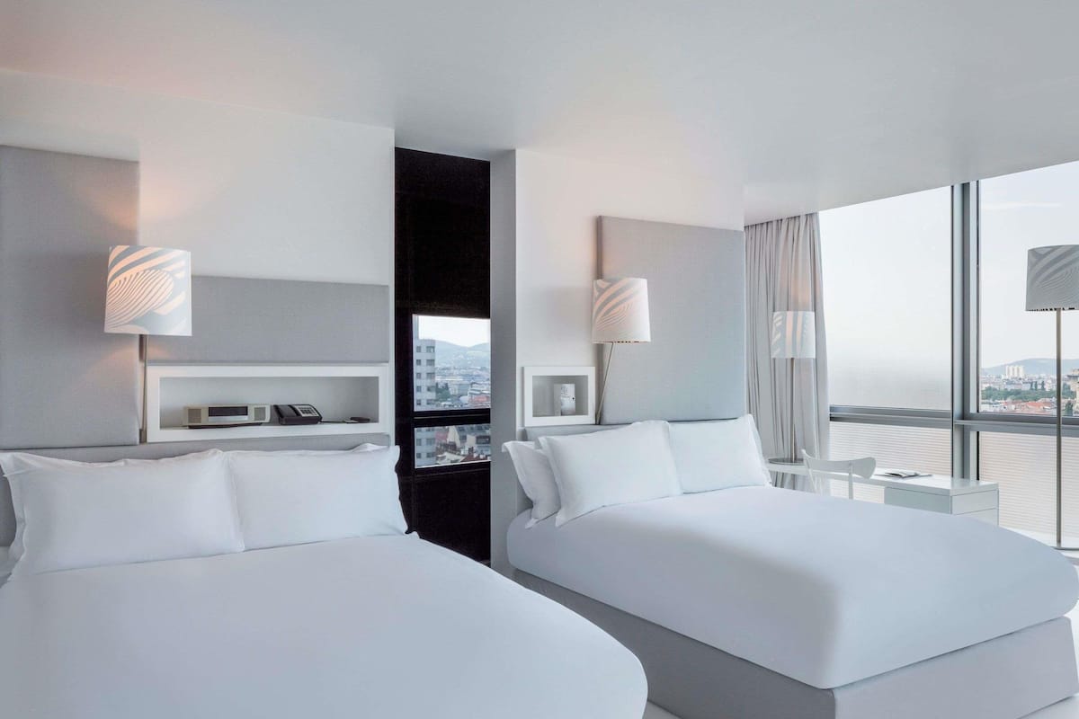 Quarto de hotel moderno com duas camas, uma pequena mesa de cabeceira, prateleira fixa com telefone e abajur, e uma grande janela com vista para a cidade.