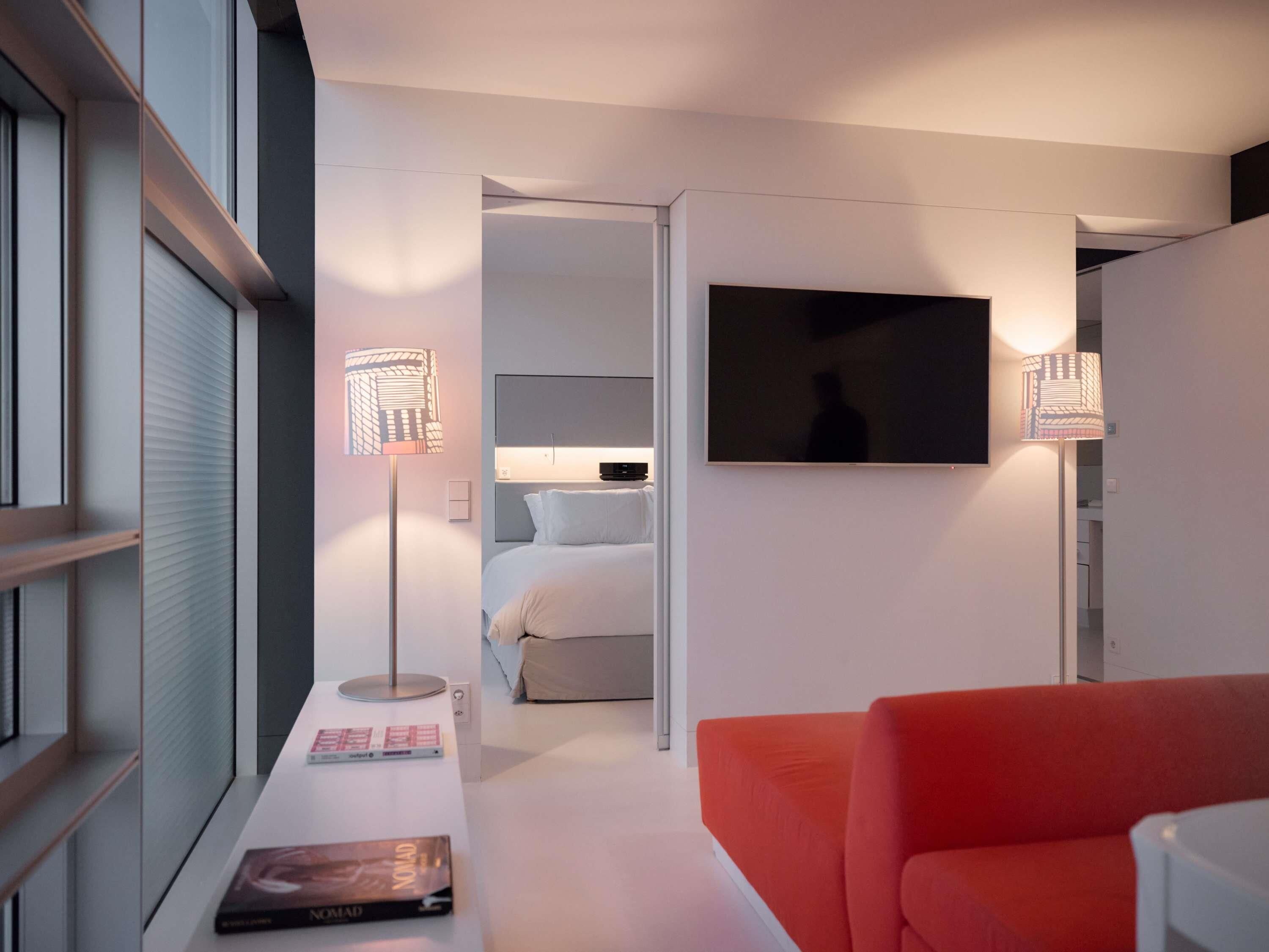 A modern hotel room with a bed, a TV mounted on the wall, a red sofa, and a desk with a lamp and books.