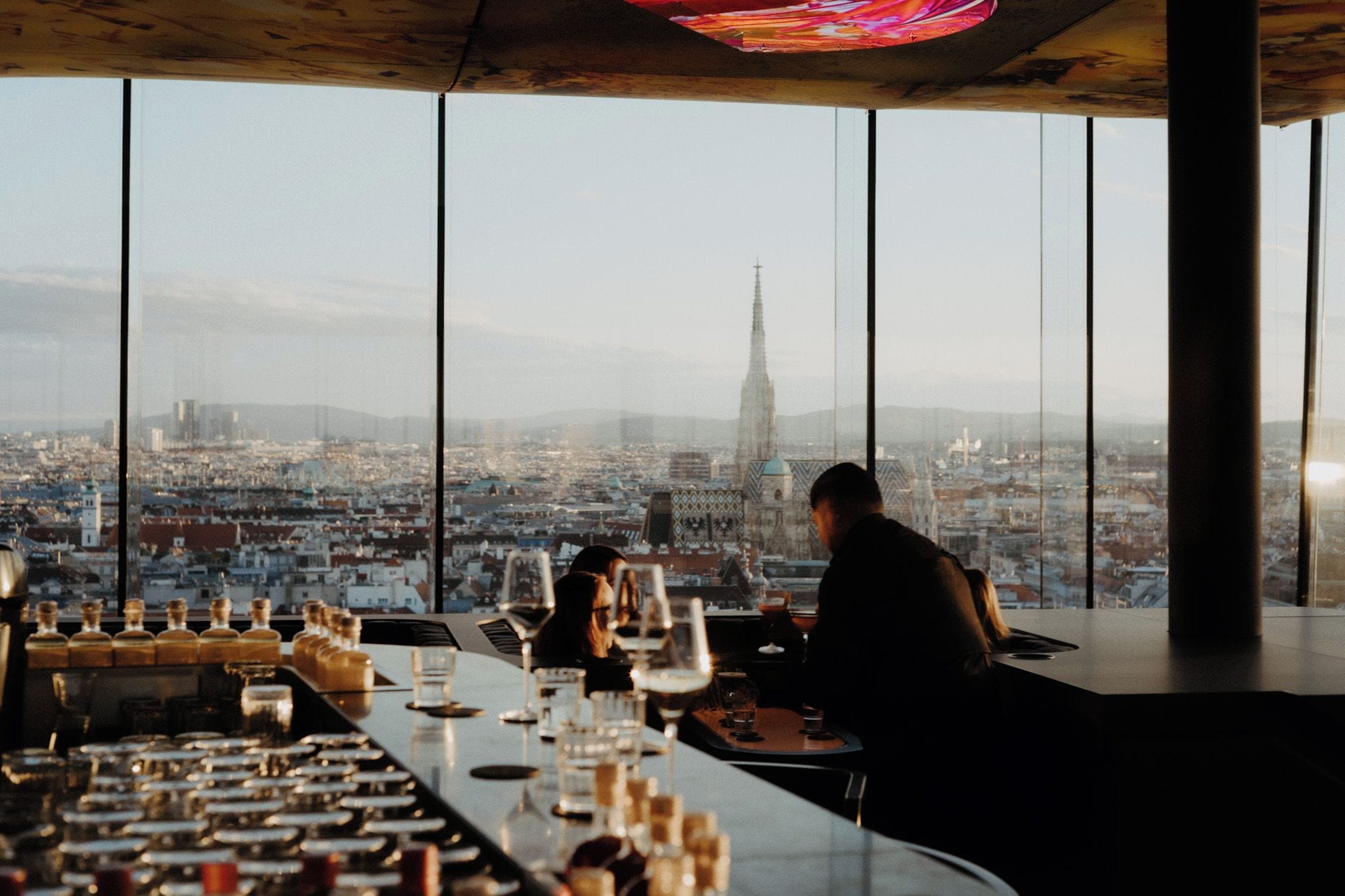 Ein Blick ins Restaurantinnere mit Stadtblick durch große Fenster, darunter ein hoher Gebäudebau in der Ferne.