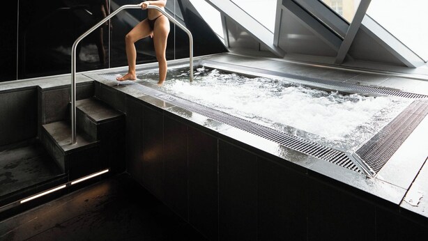 Una piscina interior moderna con paredes de vidrio transparente, una persona sentada al borde y vistas de edificios exteriores.