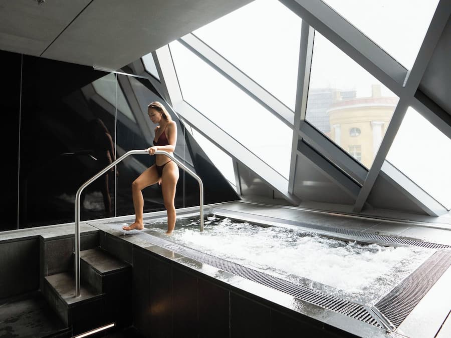 A modern indoor pool with clear glass walls, a person sitting on the edge, and a view of buildings outside.