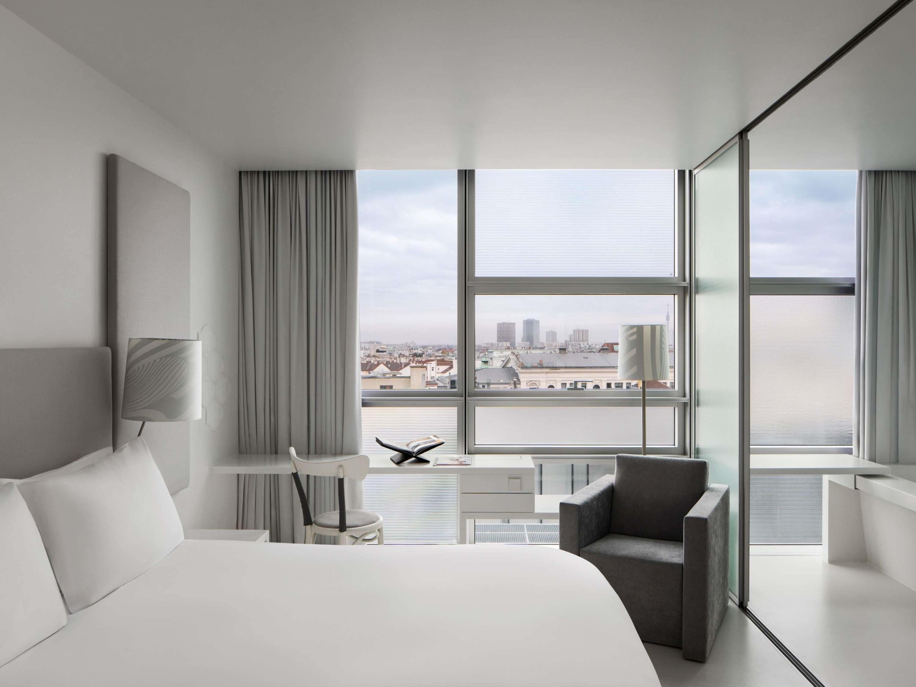 Quarto de hotel moderno com uma cama grande, uma escrivaninha, uma cadeira e vista para a cidade através de amplas janelas.