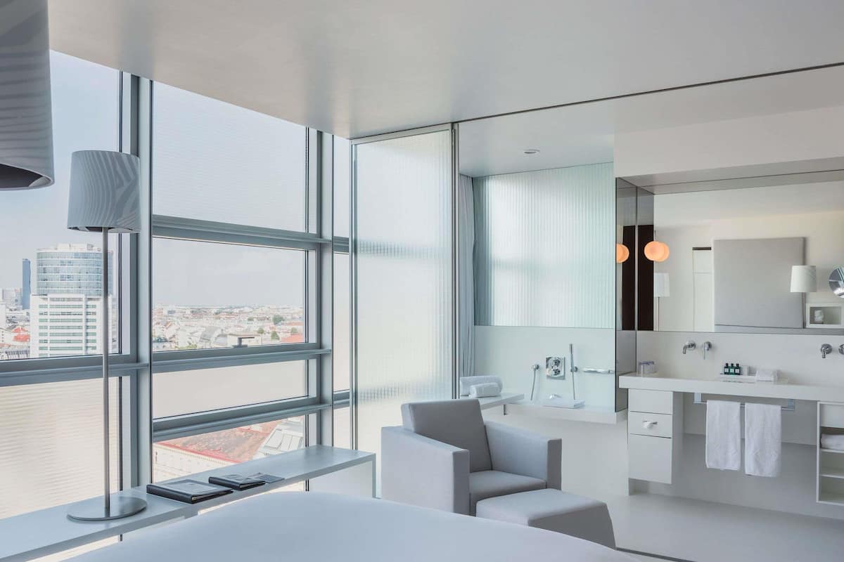 Ein modernes Hotelzimmer mit großem Fenster, einem Bett, einem Schreibtisch, einem Sessel und einem Badezimmer mit Spiegel, sowie Blick auf die Stadt.