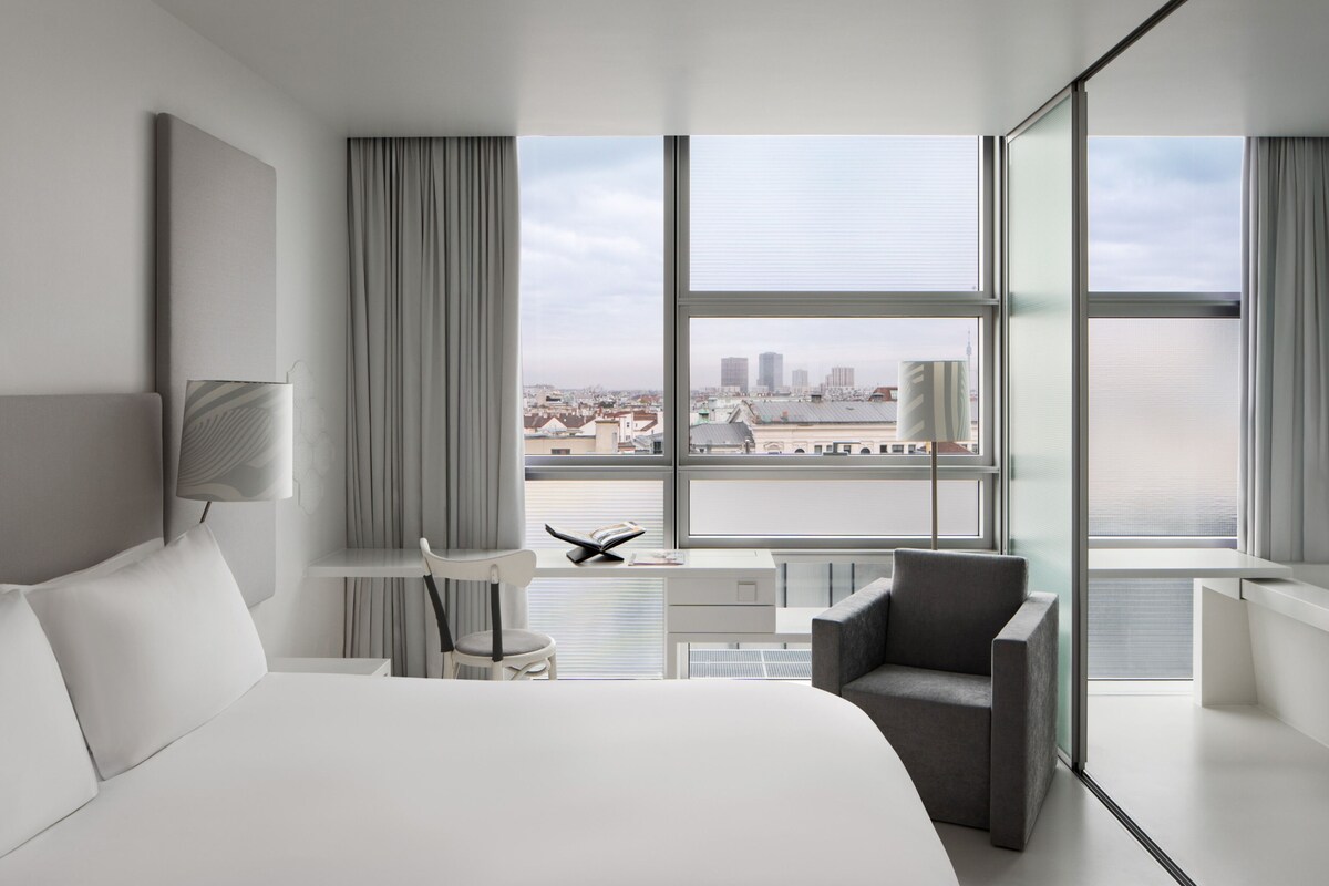 Habitación de hotel moderna con una cama grande, un escritorio, una silla y vistas de la ciudad a través de amplias ventanas.
