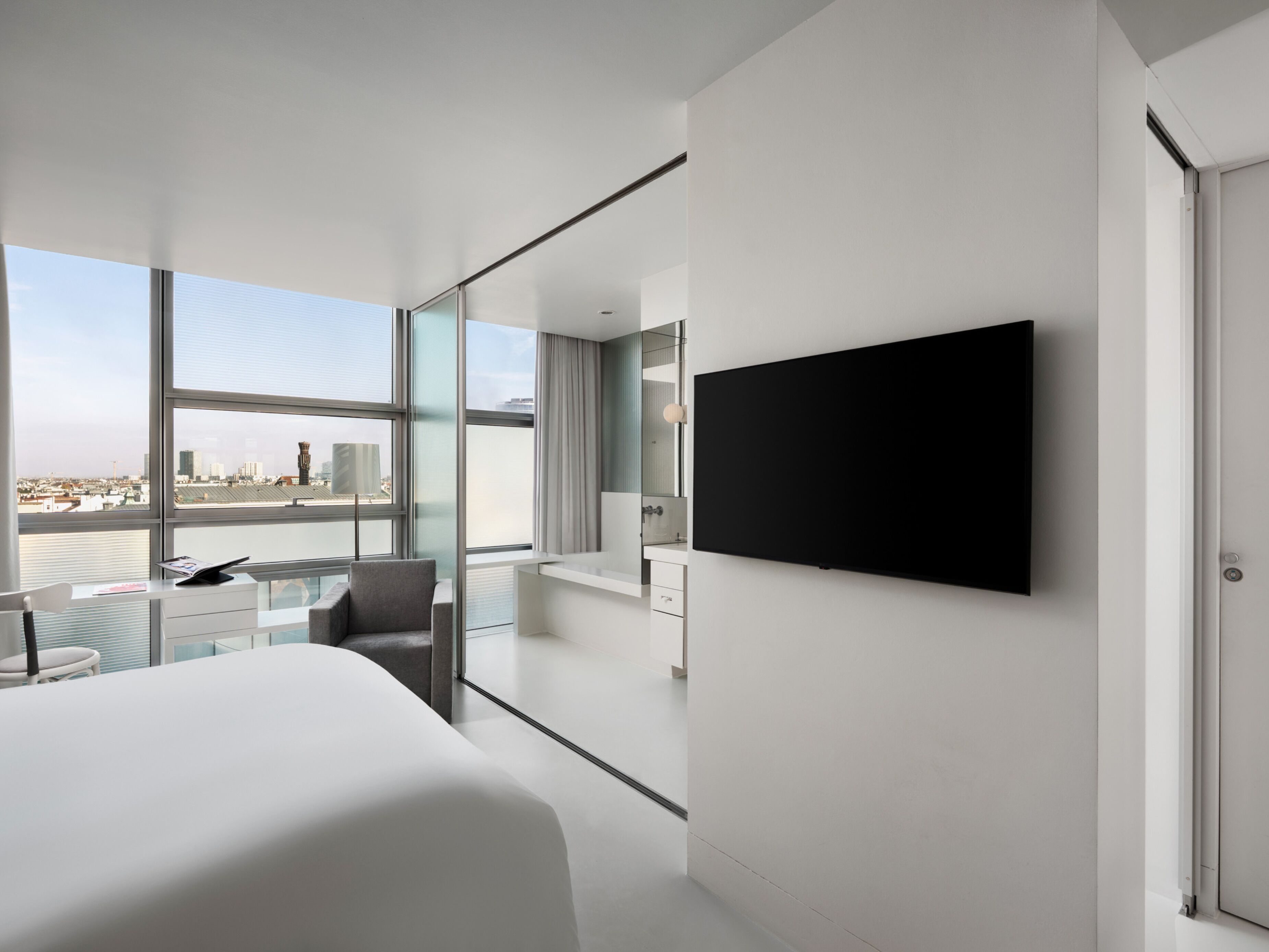 A modern hotel room with a large bed, a flat-screen TV mounted on the wall, a glass partition, and a city view.