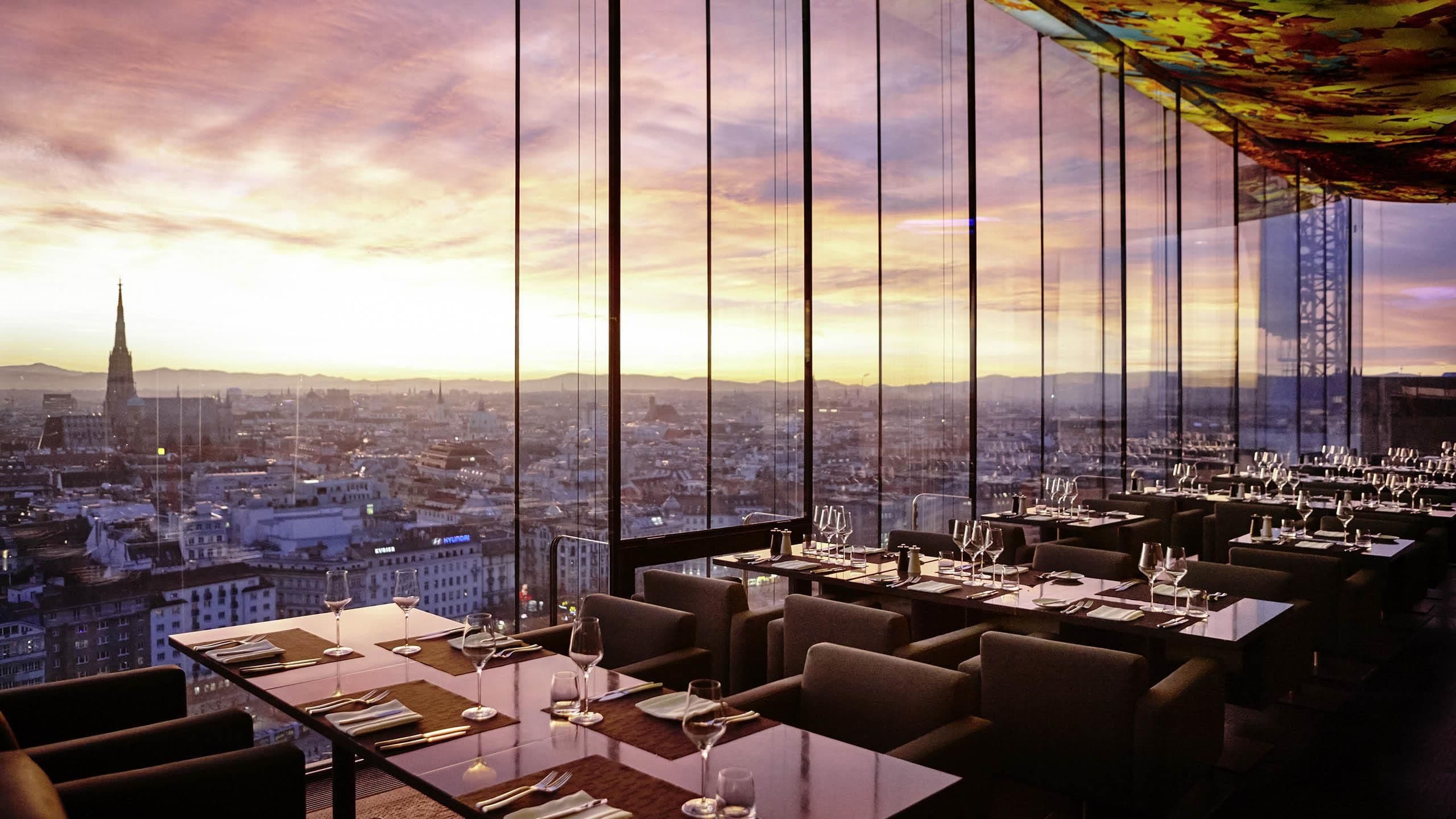 Restaurante com mesas preparadas para refeições, amplas janelas com vista para a cidade e teto alto com detalhe decorativo.