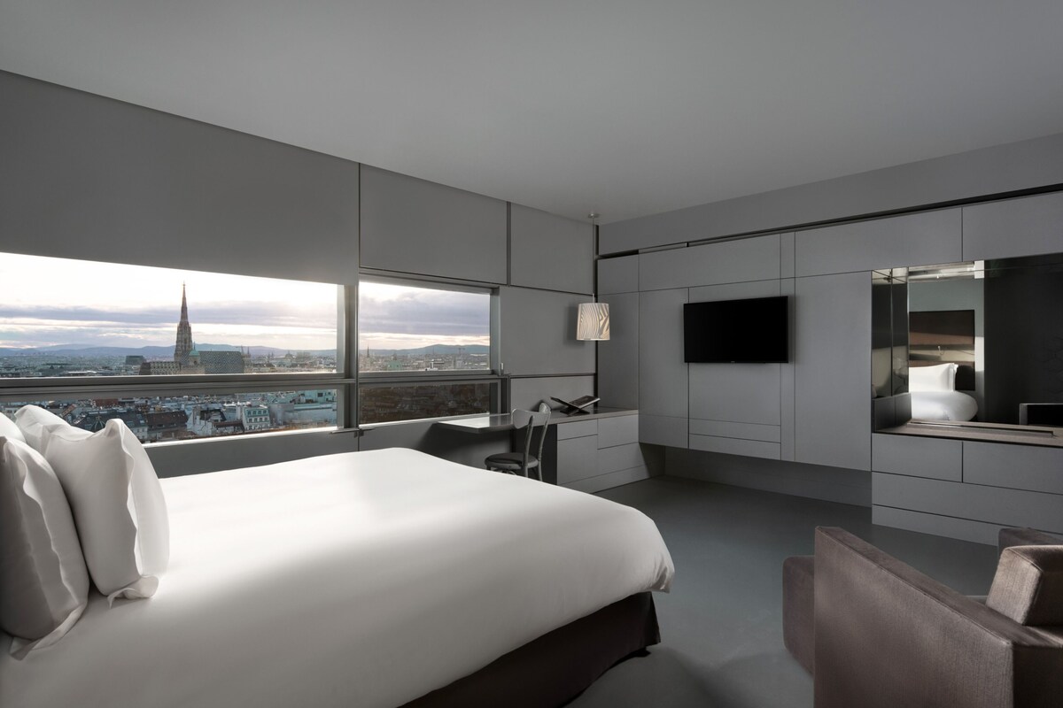 Habitación de hotel moderna con una cama grande, un escritorio, una silla, un televisor y vistas a la ciudad.