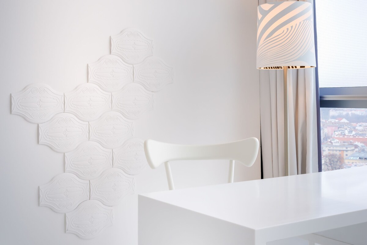 Una habitación moderna con una mesa y sillas blancas, un diseño geométrico en la pared y una ventana con cortinas.