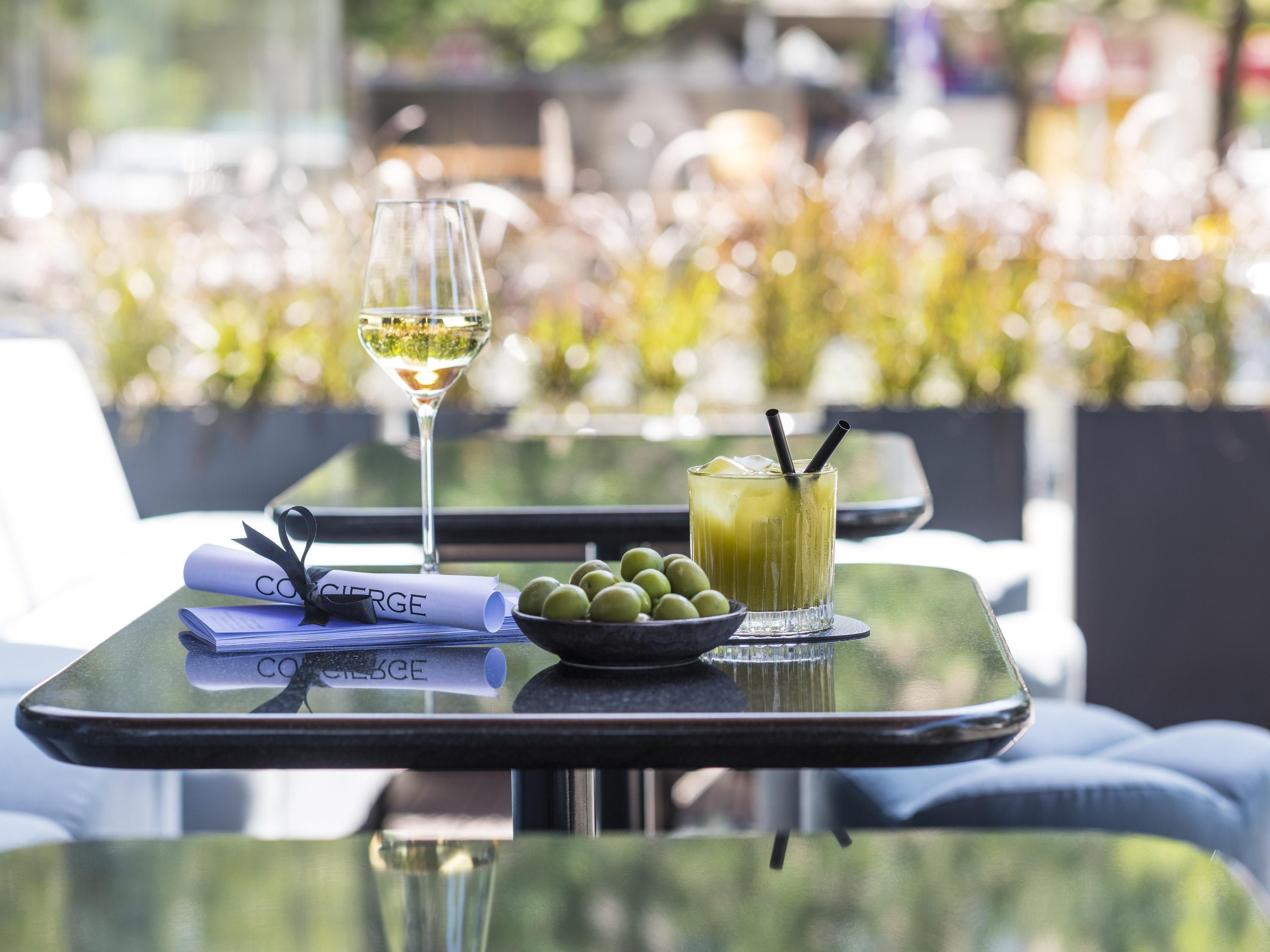 Un tavolo con un bicchiere di vino bianco, una bevanda verde con una cannuccia, una ciotola di olive e un menu arrotolato.
