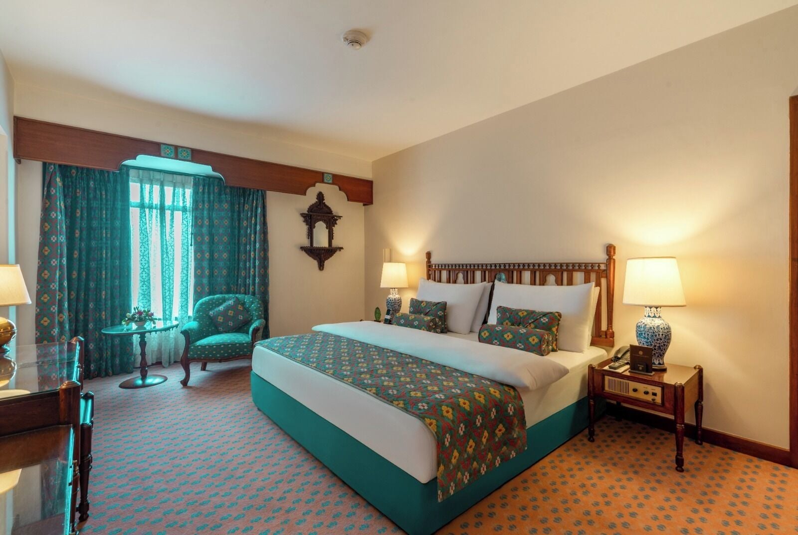 A hotel room with a large bed, two bedside tables with lamps, a chair, a small table with a vase, and a window with curtains.