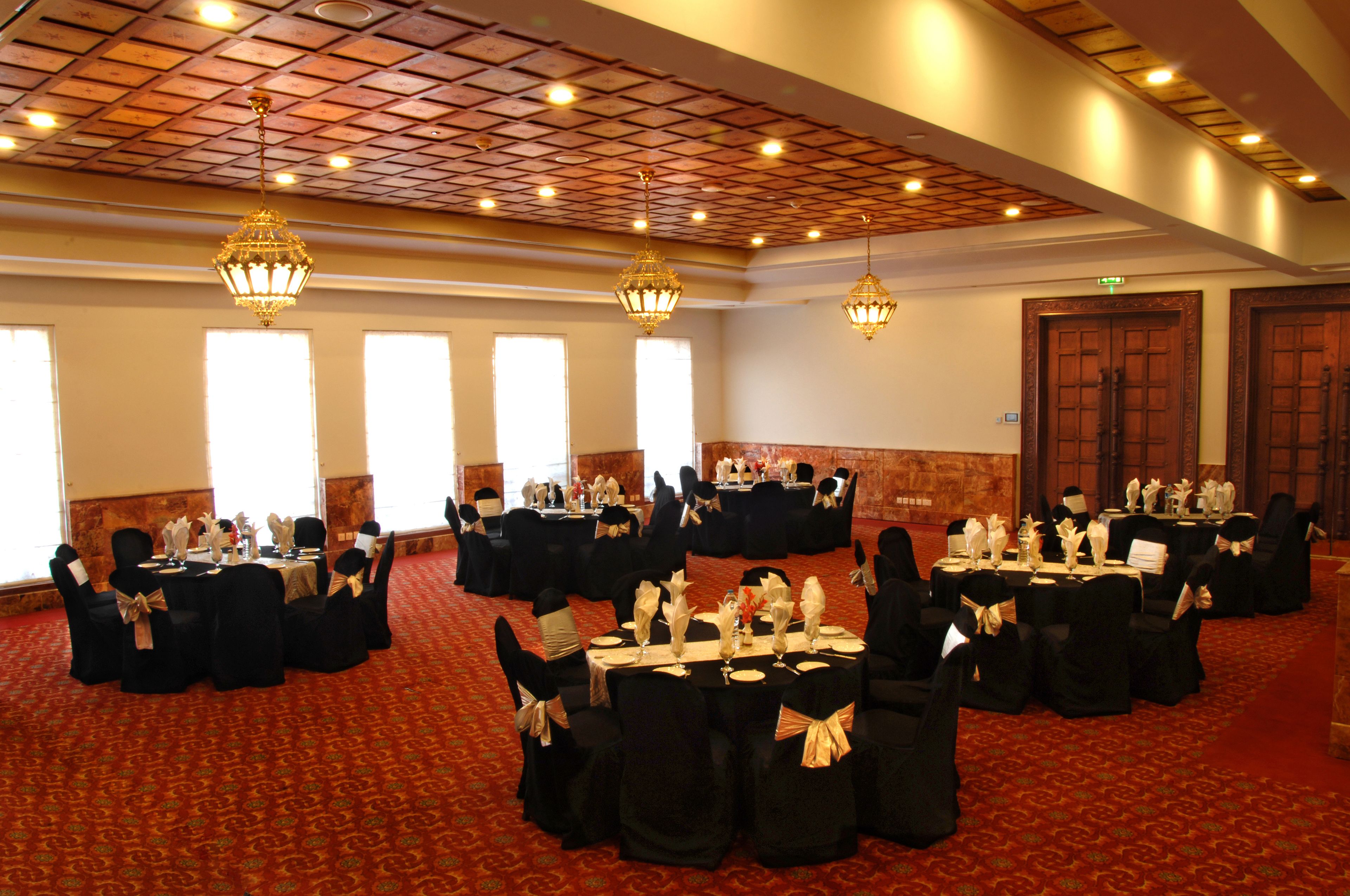 Un salón de banquetes con varias mesas redondas dispuestas para eventos, con manteles negros y sillas adornadas con cintas doradas.