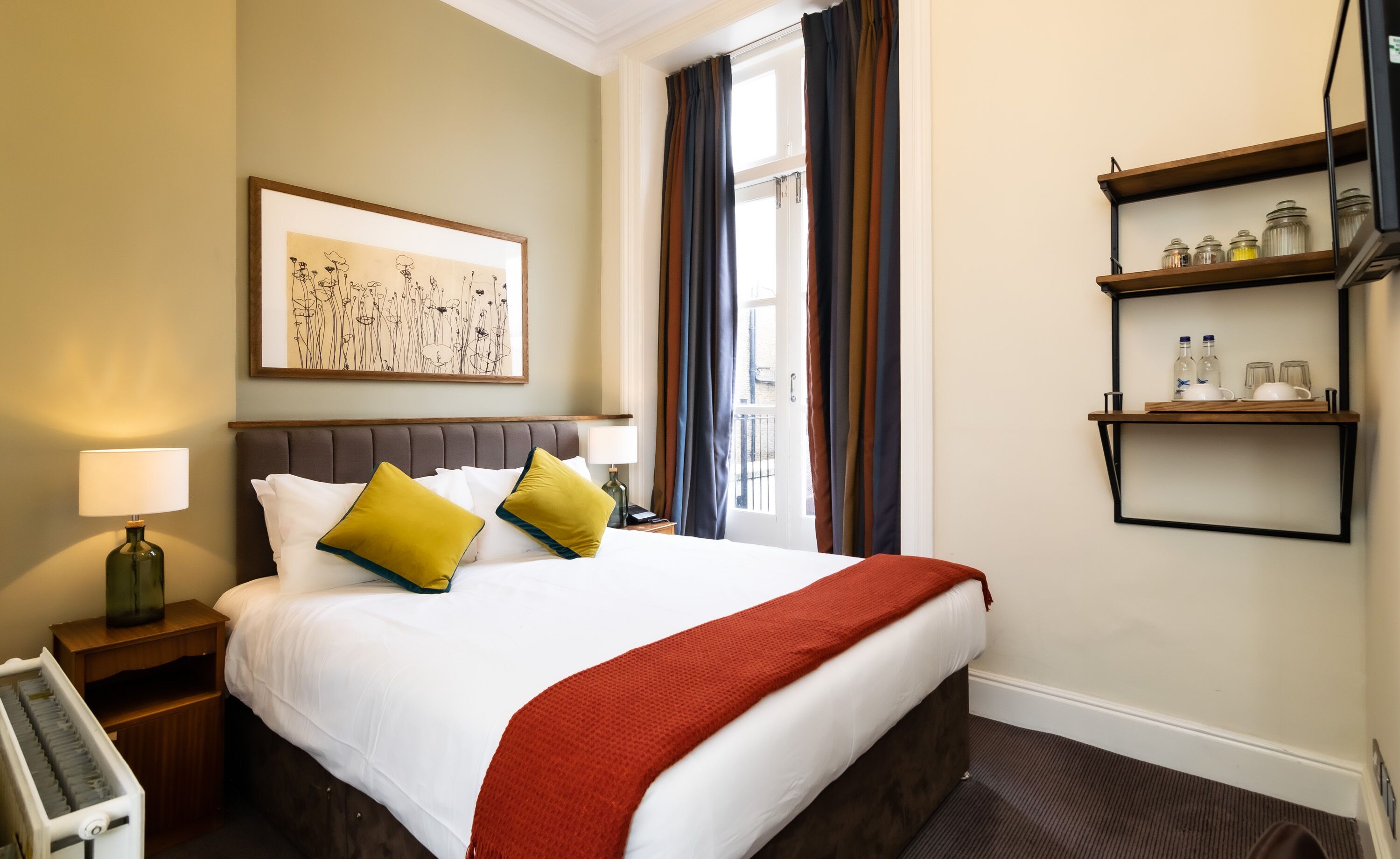A hotel room with a large bed, a bedside table with a lamp, a framed artwork on the wall, a window with curtains, and a shelf with bottles and glasses.