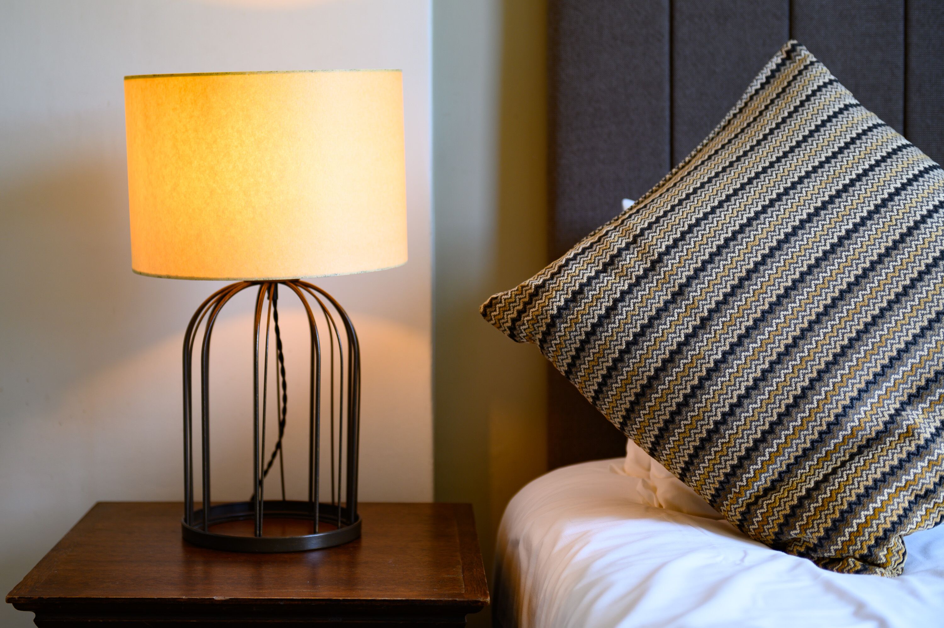 A bed with a patterned pillow and a table lamp with a cylindrical shade.
