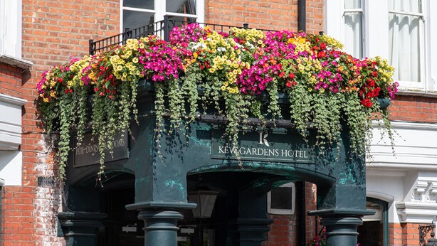 Entrada de un hotel con un letrero que dice "KEW GARDENS HOTEL" y una cesta de flores sobre la puerta. Hay dos carteles en la acera que anuncian "FIZZ FRIDAY" y "RESERVA AHORA ASADO TODOS LOS DOMINGOS".