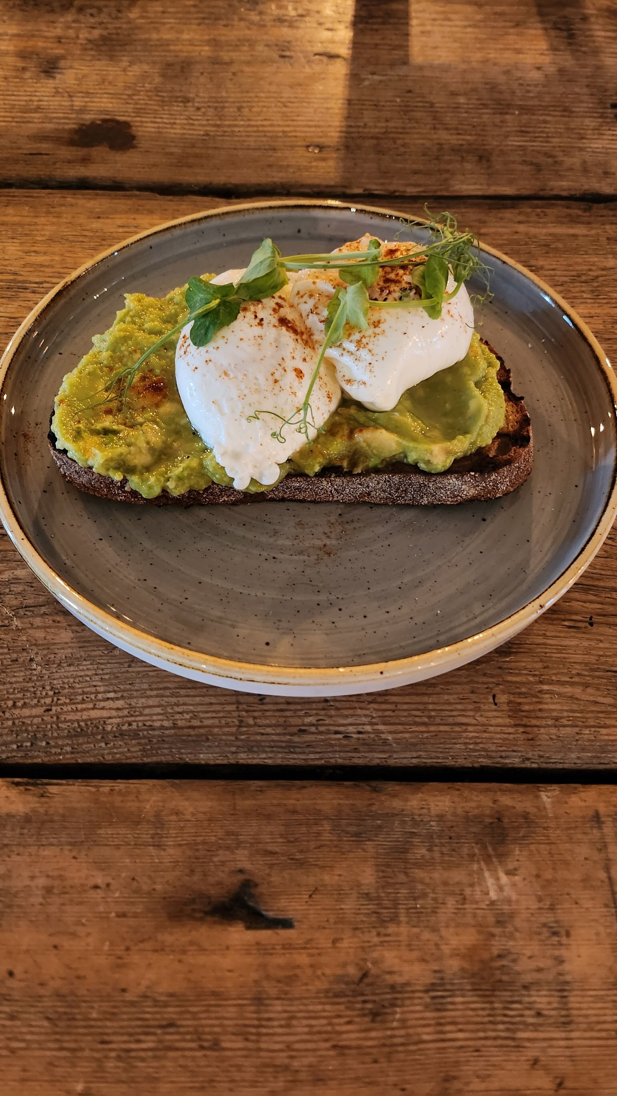 Une assiette contenant une tranche de pain grillé garnie d’avocat et surmontée d’un œuf poché, le tout décoré d’herbes, posée sur une table en bois.