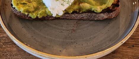 Une assiette contenant une tranche de pain grillé garnie d’avocat et surmontée d’un œuf poché, le tout décoré d’herbes, posée sur une table en bois.