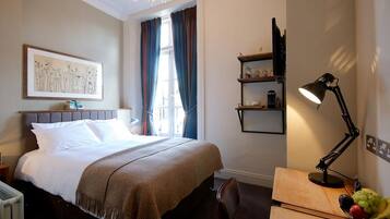 A hotel room with a bed, a desk with a lamp, a chair, a fruit bowl, a shelf with decorative items, and a window with curtains.