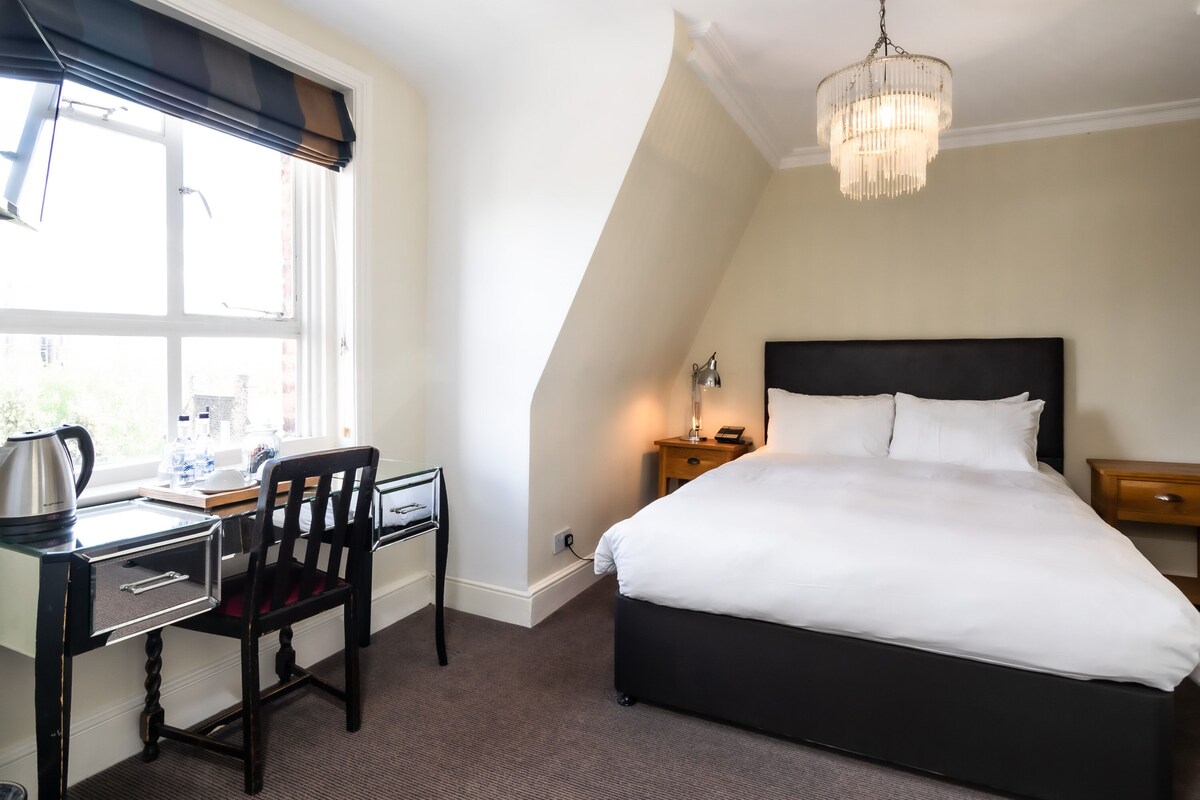 A bedroom with a bed, a desk, a chair, a nightstand, and a chandelier.