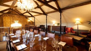 Comedor con una larga mesa de madera dispuesta para una comida, rodeada de sillas de cuero. Un candelabro cuelga del techo, y se aprecia una pared de ladrillo al fondo.