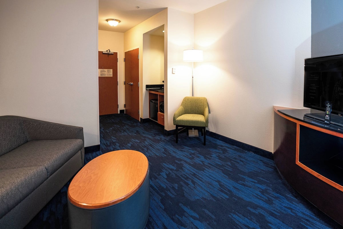 A hotel room with a gray sofa, a wooden ottoman, a green armchair, a desk with a television, and a blue carpet.