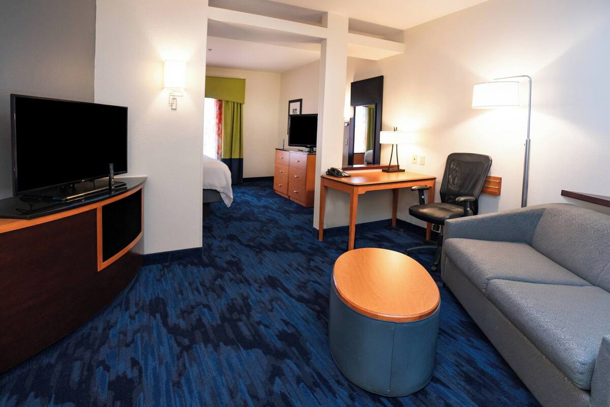A hotel room with a flat-screen TV, a desk, a chair, a sofa, and a round ottoman.