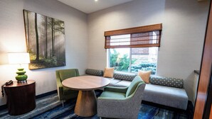 A room with a round wooden table, green armchairs, a sofa with patterned cushions, a large forest-themed painting, and a window with a view of greenery.