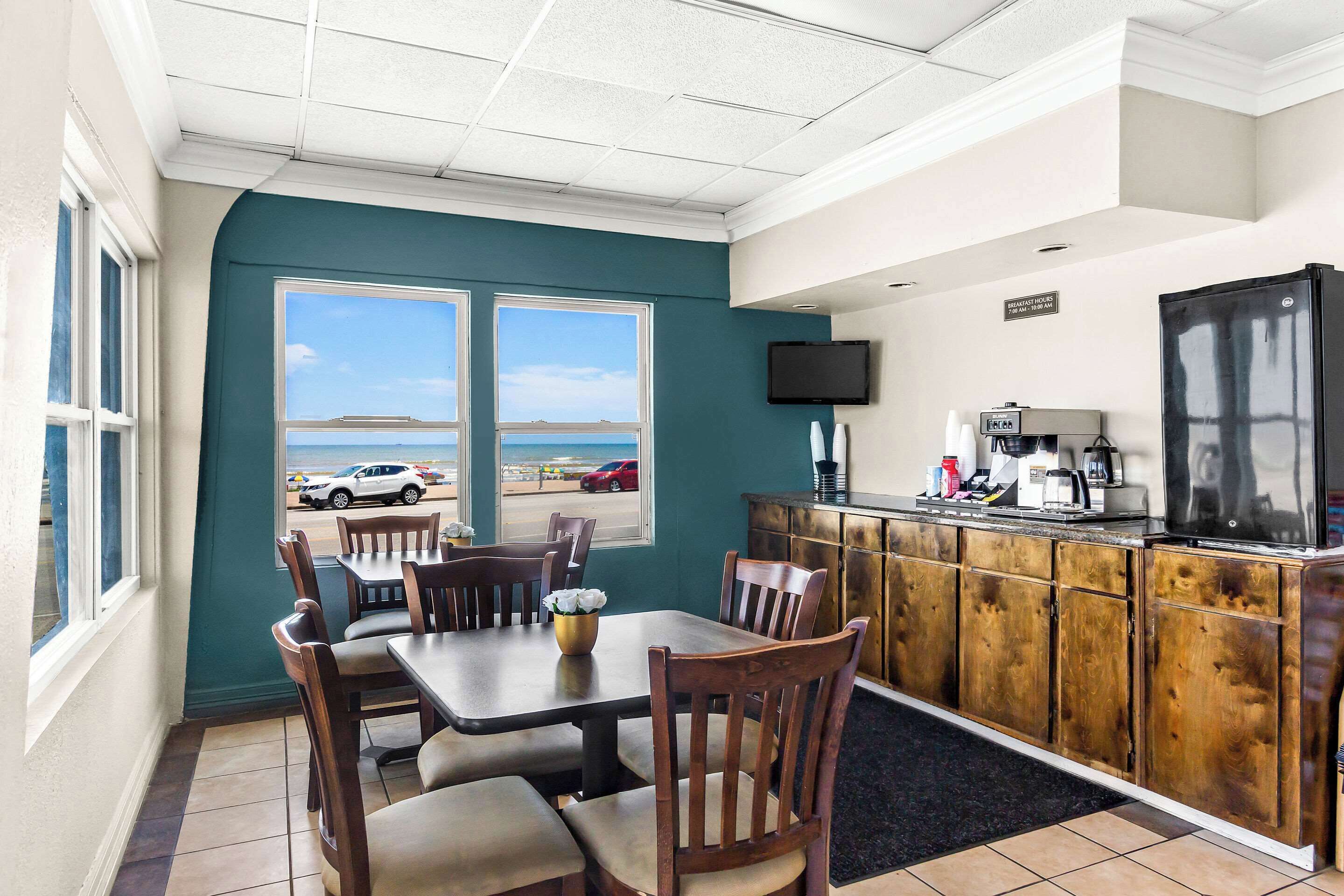 Un restaurante con vistas a la playa, mesas y sillas de madera, una zona de cocina con refrigerador y cafetera, y un televisor de pantalla plana montado en la pared.