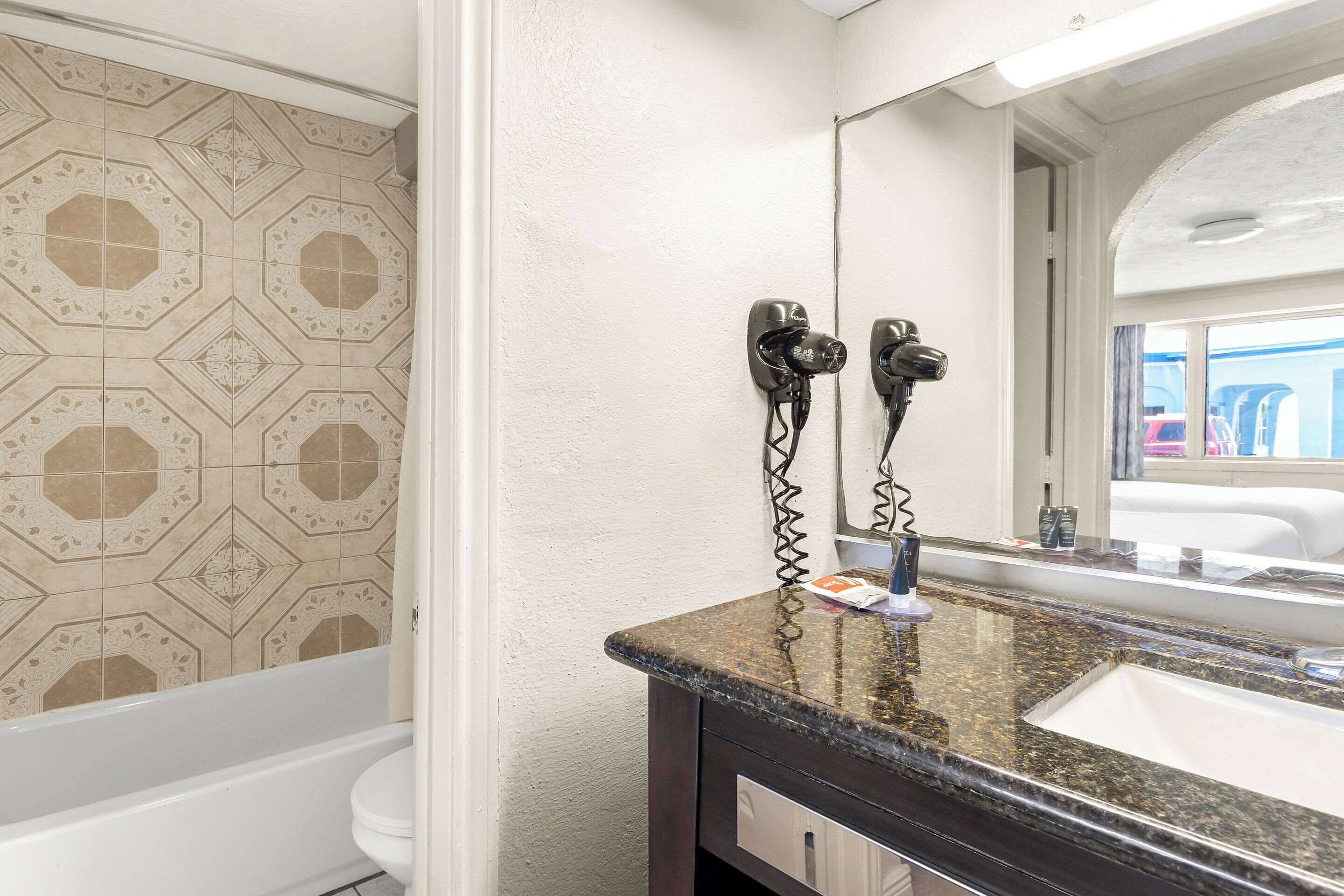 Un baño con pared de azulejos estampados, una bañera blanca, un lavamanos de granito oscuro y un espejo que refleja la vista de una ventana.