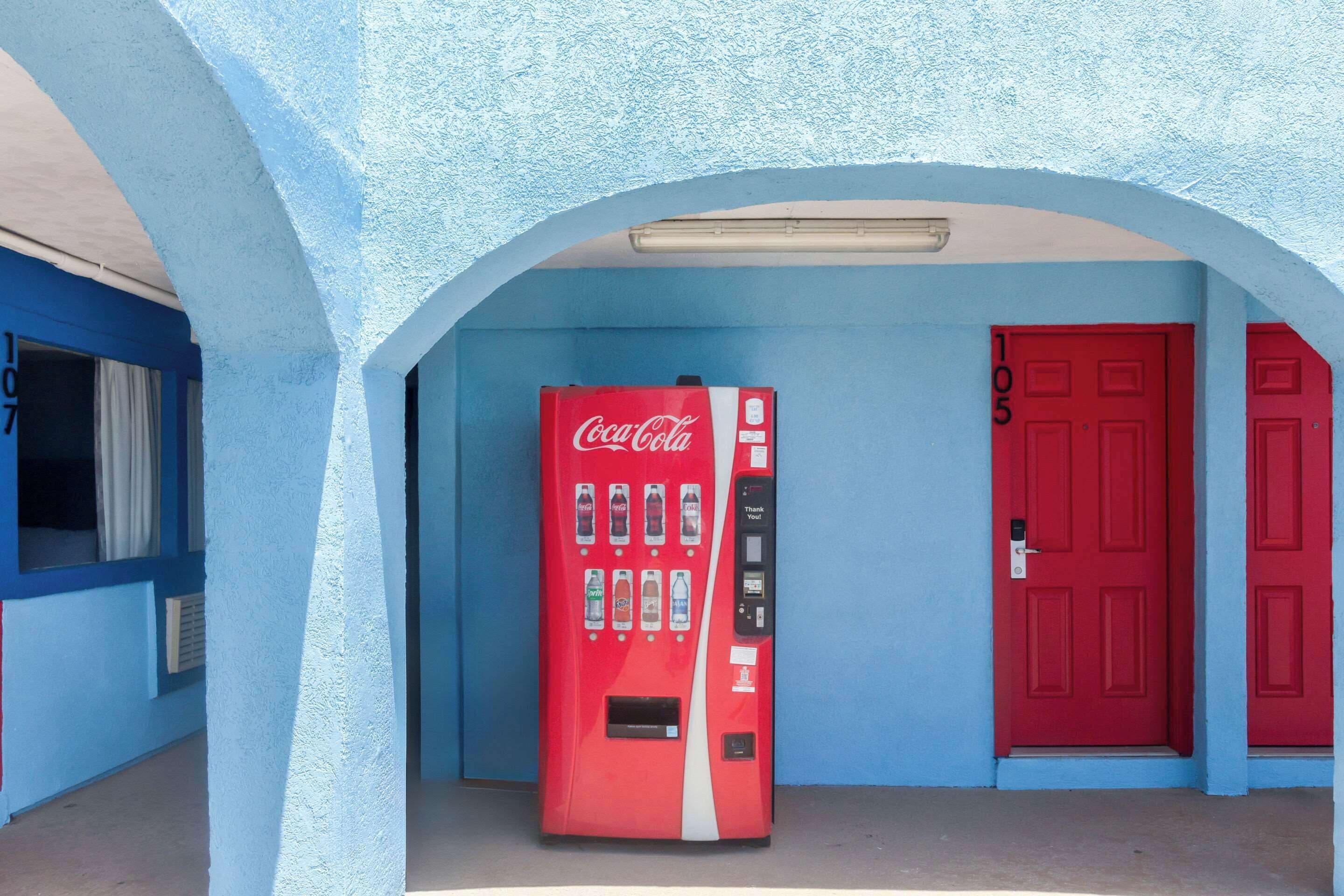 En rød Coca-Cola automt ved en væg i blå og en rød dør.