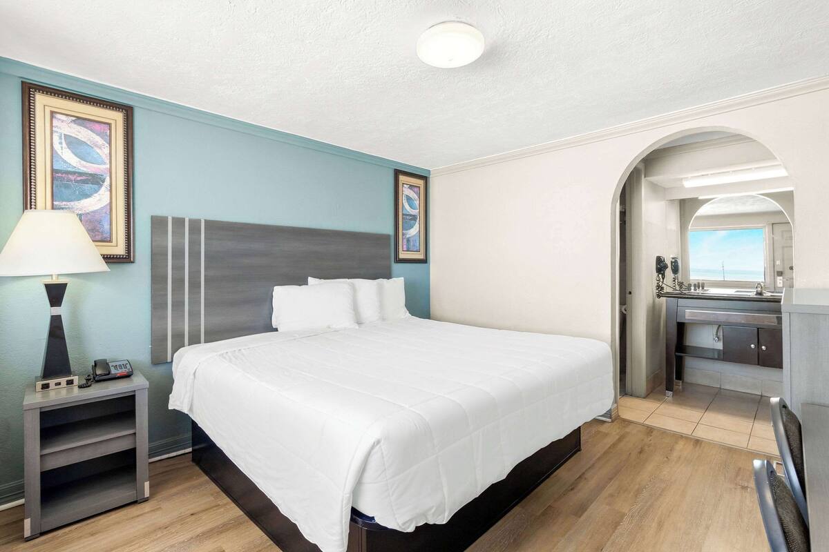 A neatly arranged hotel room with a large bed, bedside table with a lamp, and a view of the bathroom through an archway.