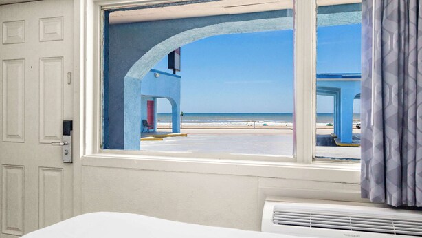 Una habitación con un ventanal grande con vistas a una playa, una puerta blanca y un equipo de aire acondicionado visible.