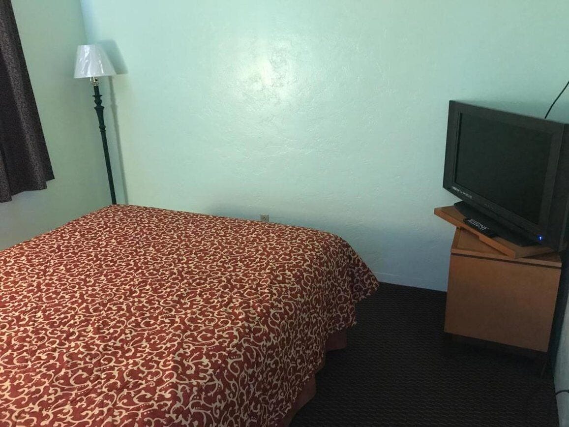 Une chambre d’hôtel avec un lit, un téléviseur posé sur un meuble en bois, une lampe et un mur sobre.