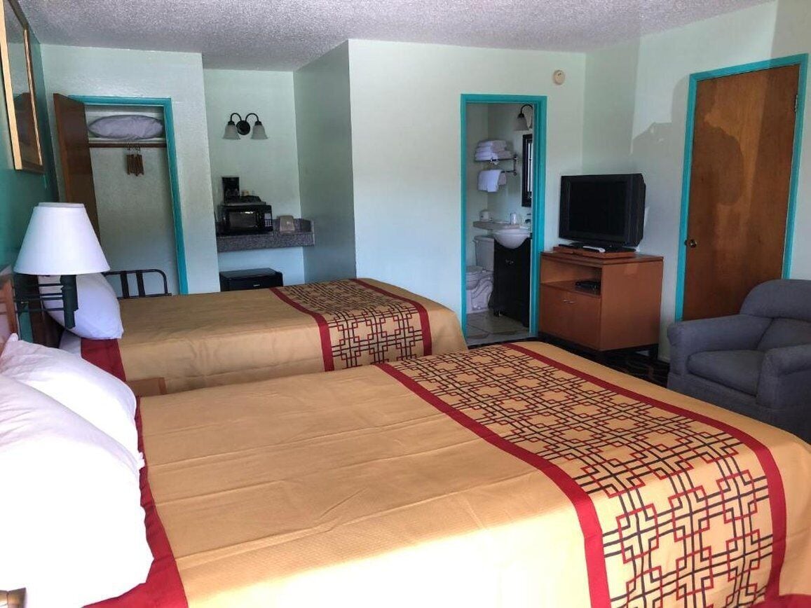 A hotel room with two beds, a desk with a television, a chair, a lamp, and a bathroom visible through an open door.