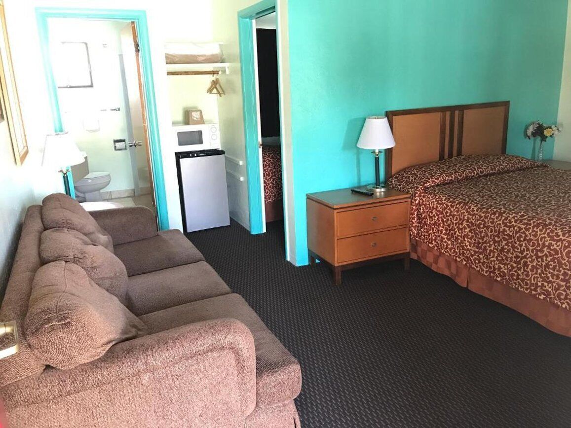 Et hotelværelse med seng, sofa, natbord, lampe, skrivebord med computer og et lille tekøkken.