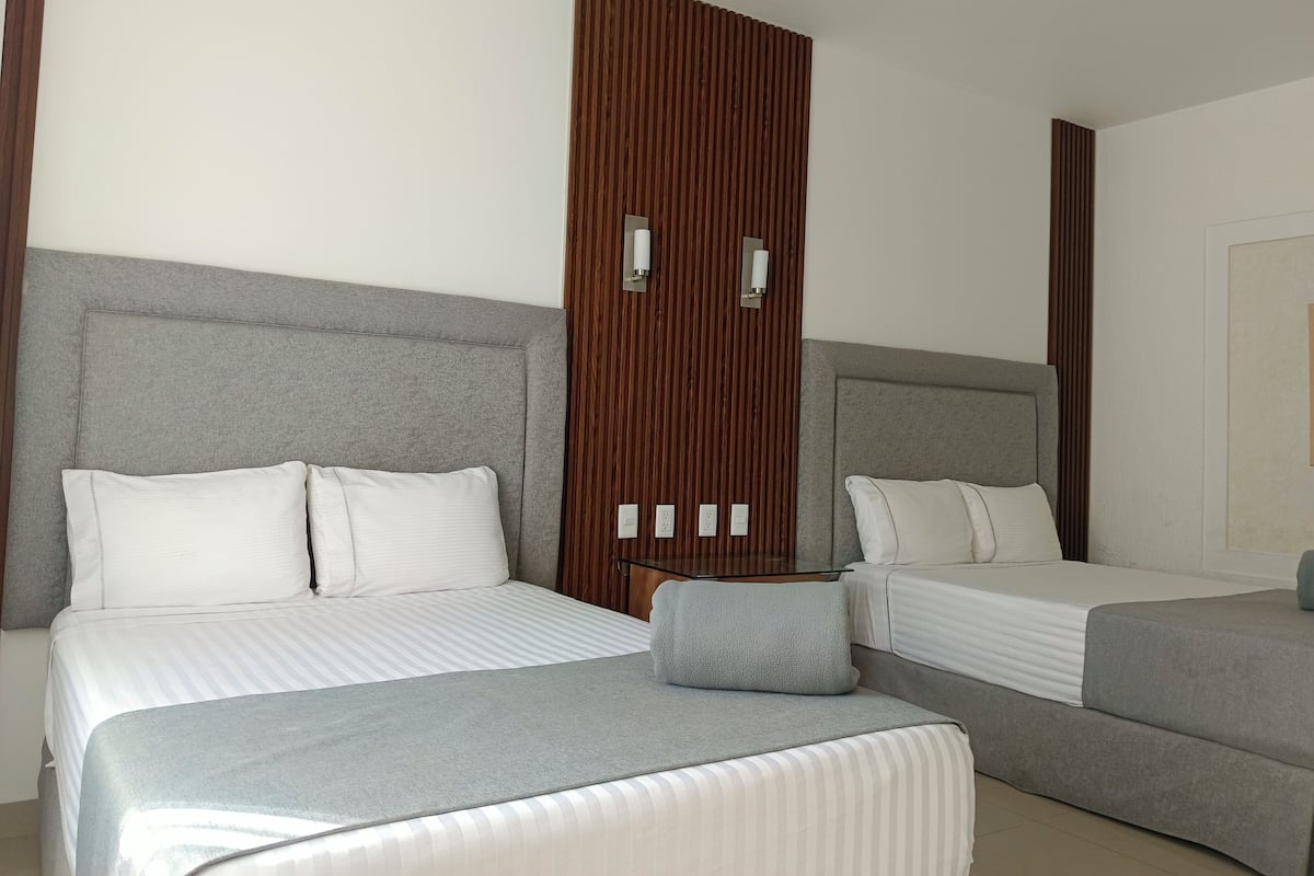 Habitación de hotel con dos camas, cada una con cabeceros grises y ropa de cama blanca. Un panel de madera divide las camas.