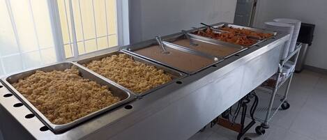 Disposição de buffet com várias bandejas de comida, uma pilha de pratos e um carrinho com recipientes.