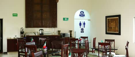 Un comedor con sillas y mesas de madera, una gran puerta de madera, una araña y un cartel que indica baños de hombres y mujeres.