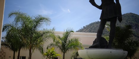 Una estatua sobre un pedestal en un patio con palmeras y un edificio al fondo.