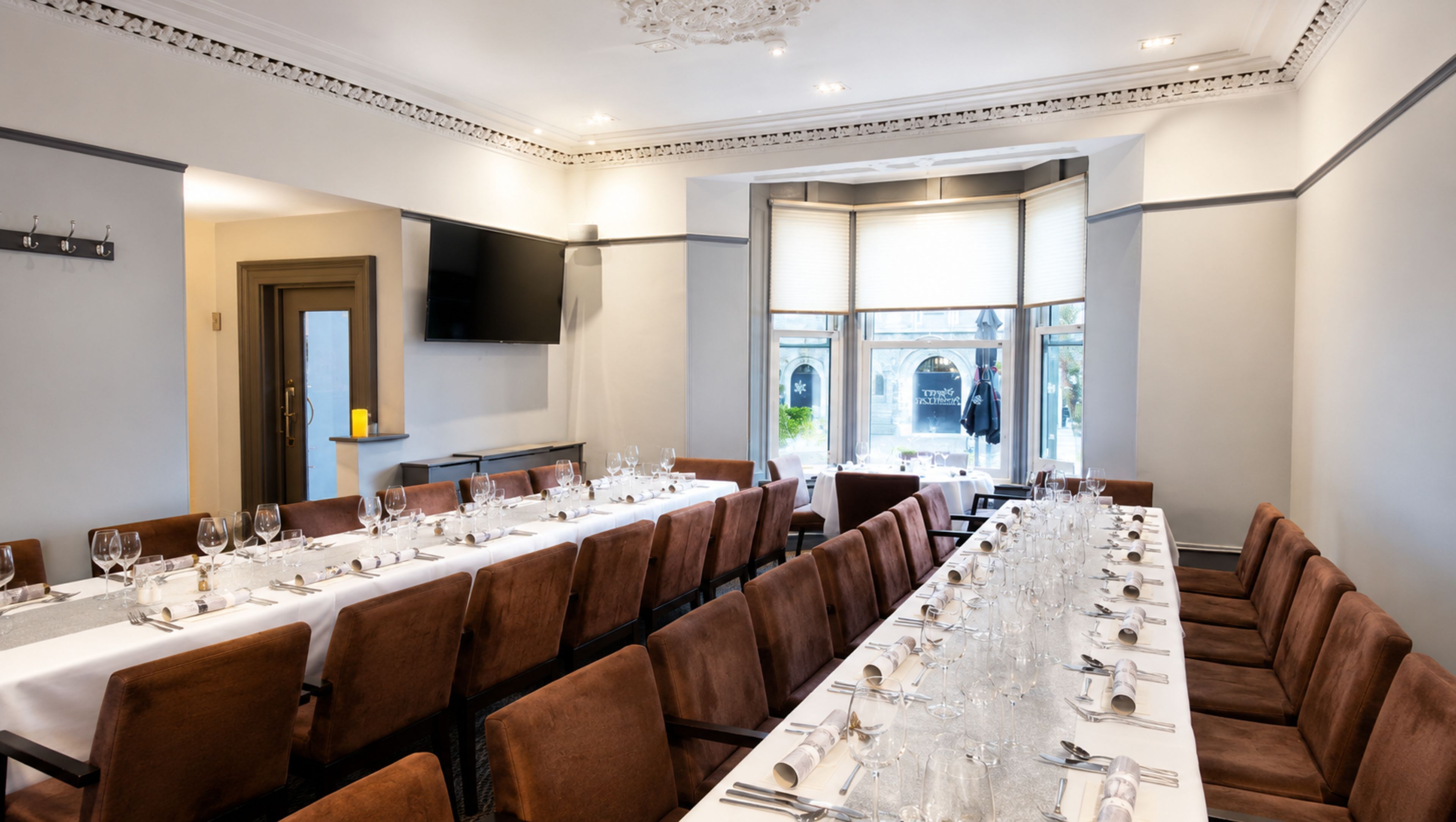 A long dining table set for a meal with multiple chairs, a flat-screen TV mounted on the wall, and a view of the outdoors through large windows.