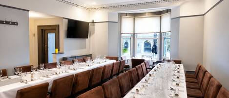 A long dining table set for a meal with multiple chairs, a flat-screen TV mounted on the wall, and a view of the outdoors through large windows.