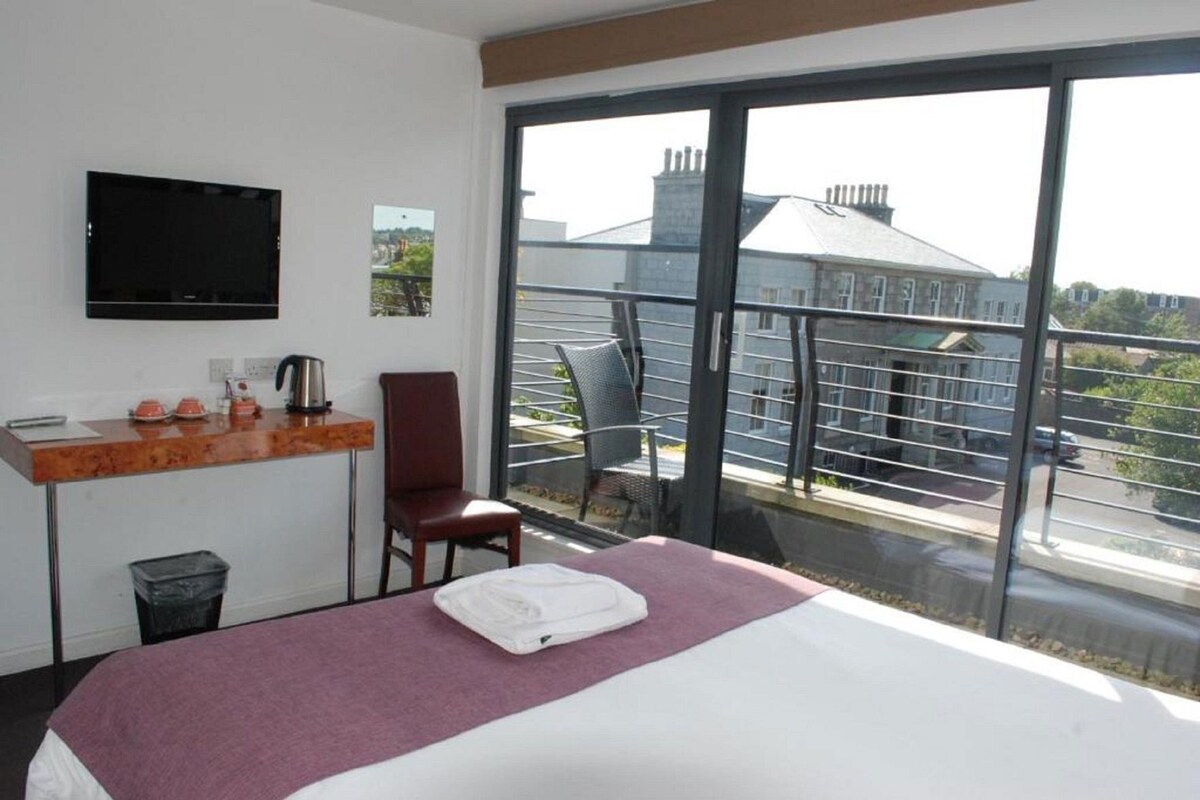 A hotel room with a bed, a desk, a chair, a TV, and a balcony with a view of buildings.