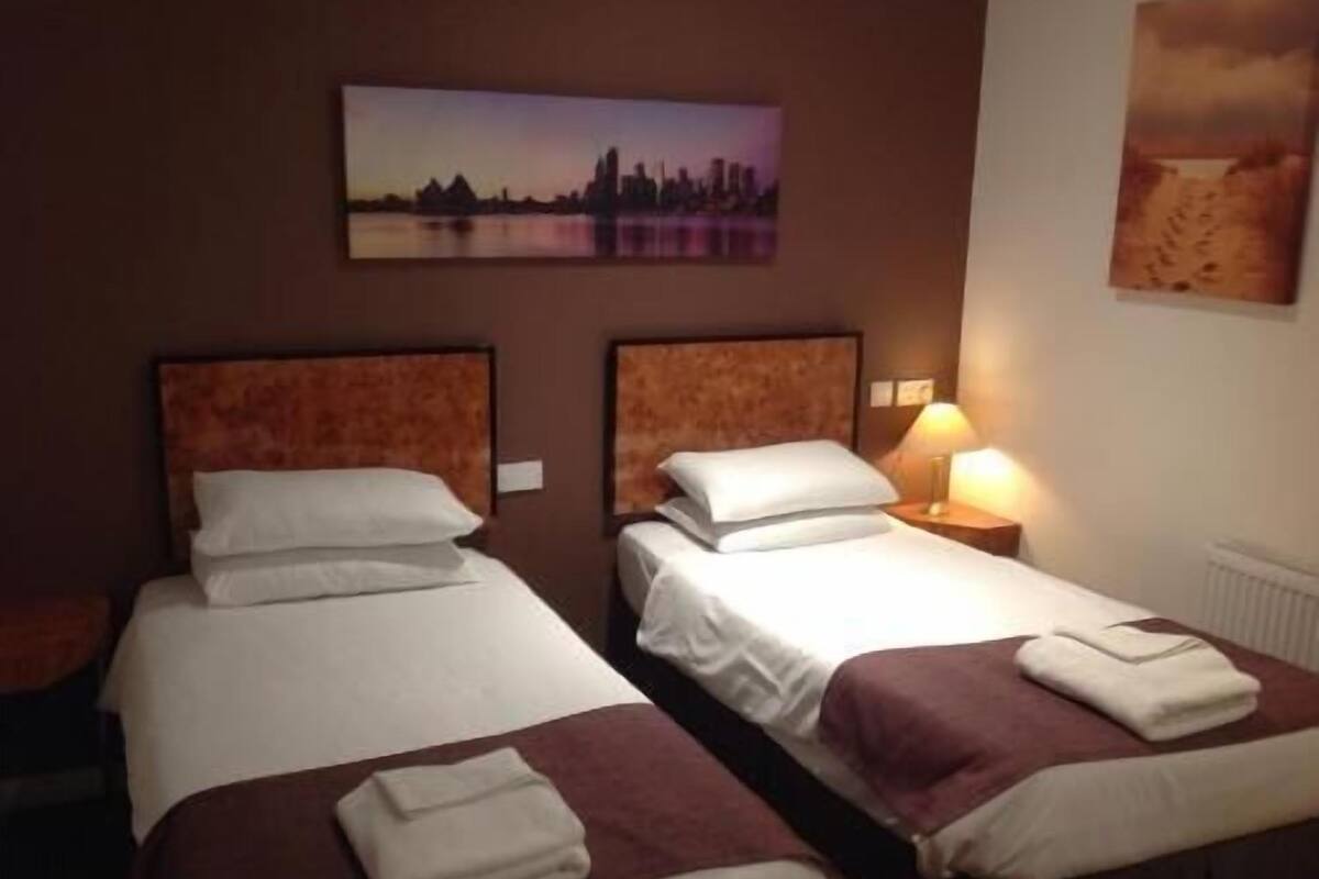 A hotel room with two beds, a cityscape painting, and a lamp.