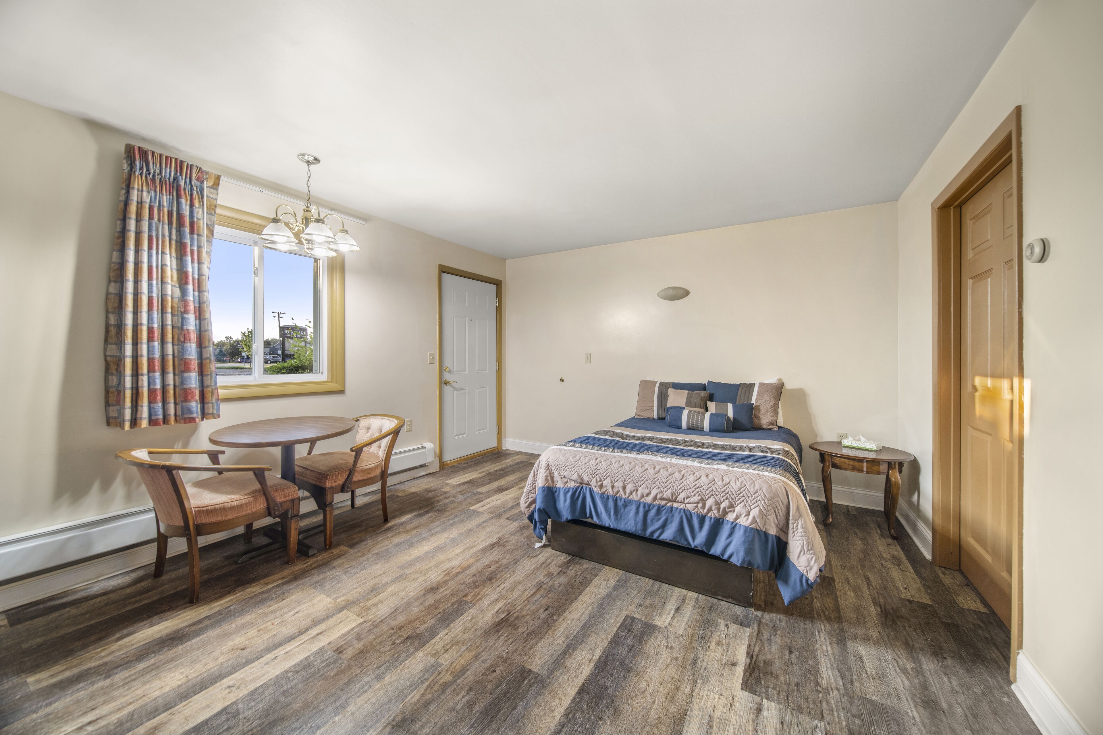 A bedroom with a bed, a round table, two chairs, a window with curtains, and a door.