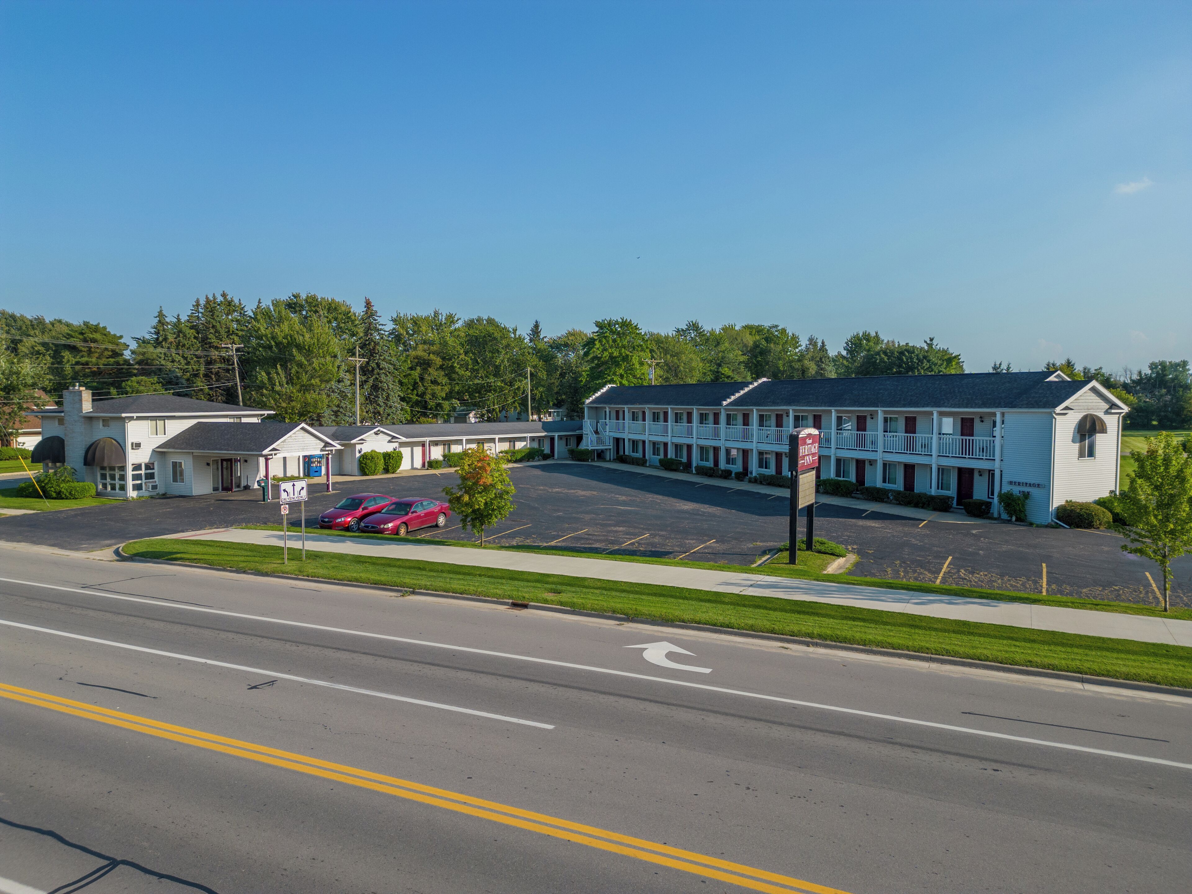 A motel with a parking lot, a road in the foreground, and trees in the background.