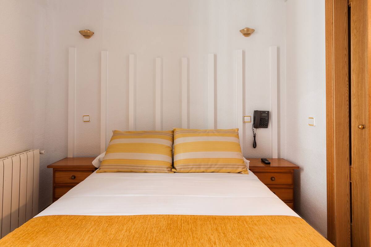 Una cama bien hecha con dos almohadas, una mesita de noche de madera con un teléfono y un radiador en la pared.